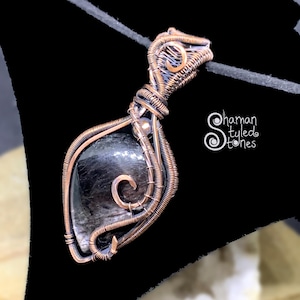 Peut inclure: Un pendentif artisanal avec une pierre sombre et polie enveloppée dans du fil de cuivre. Le travail du fil comprend des volutes complexes et une bélière pour un collier. Le pendentif est présenté sur fond noir. Le texte "Shaman Styled Stones" est visible.