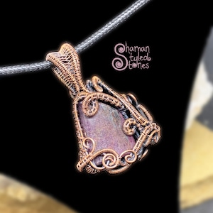 May include: A purple and grey gemstone pendant wrapped in copper wire with a decorative swirl design. The pendant is hanging from a black cord. "Shaman Styled Stones" is written in pink text on a white background.