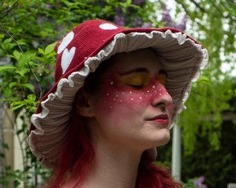 May include: A burgundy mushroom-shaped hat with white heart-shaped accents and a ruffled brim. The hat is paired with a rust-colored corset-style top and a cream-colored floral print dress. Yellow ribbons and accents add detail to the outfit.