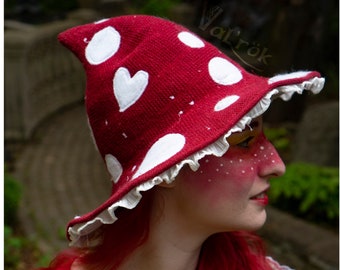 May include: A red and white mushroom-shaped hat with white polka dots and a white heart design. The hat has a white ruffle trim and is worn by a person with red hair.
