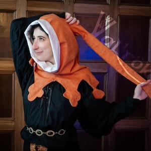May include: An orange and white medieval hood with long, pointed tails. The hood is worn over a black tunic with a decorative belt. The hood's design features a scalloped edge and a white interior lining.
