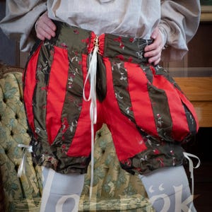 May include: Puffy shorts featuring alternating vertical panels of solid red fabric and dark olive green fabric with a delicate floral pattern. The shorts have a high waistband with a central white ribbon lace-up closure and white ribbon ties at the leg cuffs.