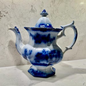 May include: A ceramic teapot featuring a white base with a deep blue, cloud-like mottled pattern. The teapot has a faceted, hexagonal body, a curved handle, a pointed spout, and a matching lid with a small, pointed finial on top.