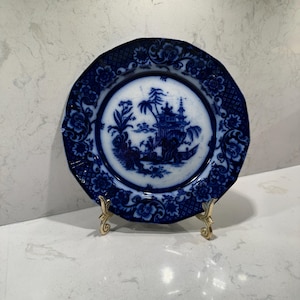 May include: A round ceramic plate featuring a dark blue and white transferware pattern. The center depicts a landscape with a pagoda and trees, while the scalloped rim is decorated with floral motifs. The plate rests on a small gold metal stand.