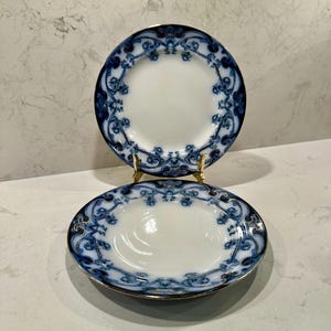 May include: Two round porcelain plates featuring a white center and a wide border decorated with intricate dark blue floral scrollwork patterns. The edges of both plates are finished with a thin gold trim, and one plate is displayed upright on a stand.