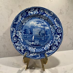 May include: A blue and white decorative plate with a landscape scene in the center. The plate has a detailed floral and scrollwork border. The plate is displayed on a gold-colored stand, showcasing its intricate design and vintage aesthetic.
