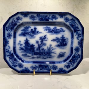 May include: An octagonal blue and white porcelain serving platter. The platter features a central landscape scene with pagodas and trees, surrounded by a floral border. The platter is supported by two small gold-colored stands.