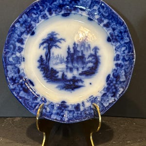 May include: Antique blue and white plate with a landscape scene. The plate features a central image of a castle and trees, surrounded by a floral border. The plate is displayed on a gold-colored stand.