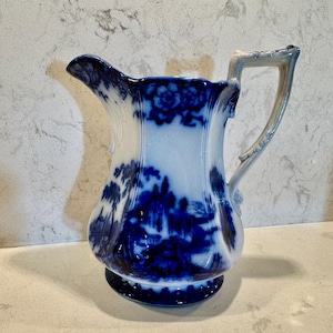 May include: A white and blue ceramic pitcher with a detailed landscape design. The pitcher features a curved spout, a decorative handle, and a round base. The blue design is set against a white background.