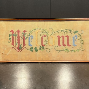 May include: A framed needlepoint artwork with the word "Welcome" stitched in colorful thread. The letters are in red, blue, yellow, and green, with decorative vines. The frame is a light brown color.