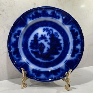 May include: A round ceramic plate featuring a deep blue and white transferware pattern. The center displays a landscape scene with trees and structures, while the scalloped rim has a decorative border. The plate rests on a gold metal stand.