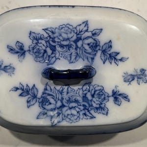 May include: A white ceramic serving dish with a blue and white floral design. The dish has a lid with a blue bamboo-style handle.