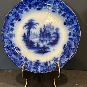 May include: Antique blue and white plate with a landscape scene. The plate features a central image of a castle and trees, surrounded by a floral border. The plate is displayed on a gold-colored stand.