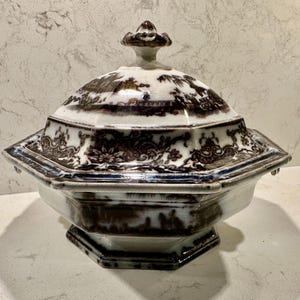 May include: An octagonal ceramic tureen with a lid, featuring a dark brown transferware pattern on a white background. The design includes intricate floral borders and landscape scenes. The lid is topped with a decorative finial handle.