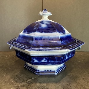 May include: A blue and white ceramic serving dish with a lid. The dish has a geometric design with a hexagonal base and a domed lid. The lid is topped with a small, decorative finial. The dish is approximately 10 inches wide and 8 inches tall.