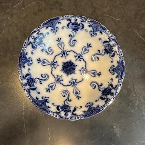 May include: A round, antique white plate with a blue floral design. The plate features a scalloped edge and intricate blue patterns, including floral motifs and swirling lines. The center of the plate has a starburst design.