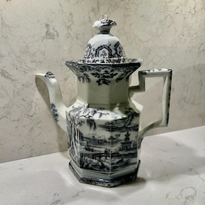 May include: A hexagonal ceramic teapot featuring a white base with intricate dark blue landscape illustrations, including pagodas, trees, and bridges. It has a matching lid with a finial, a curved spout, and a geometric handle.