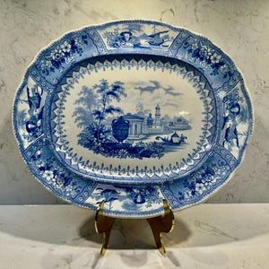 May include: An oval, antique blue and white ceramic platter with a scalloped edge. The center features a detailed landscape scene, while the border showcases floral and architectural motifs. The platter is displayed on a gold-toned stand.
