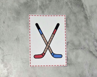 May include: A rectangular sticker with a white background and a red dashed border. The design features two crossed hockey sticks, one with a red blade and blue handle, the other with a blue blade and red handle. The text HOLLANDER and ROBANOV is visible.