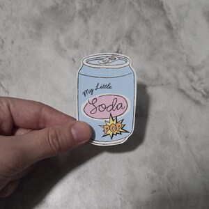 May include: A sticker featuring a cartoon soda can design. The can is light blue with the words "my Little Soda" in cursive and a pink oval with the word "Soda" inside. A yellow burst with the word "POP" is at the bottom.