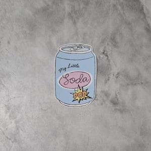 May include: A light blue sticker in the shape of a soda can with the words "my Little Soda" in cursive and a pink oval. A yellow "POP" burst graphic is at the bottom.