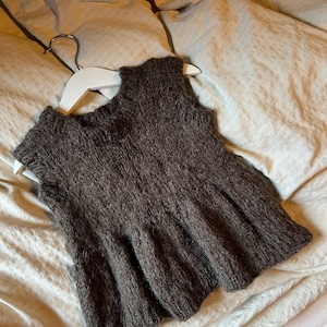 Puede incluir: Un top sin mangas de punto marrón con dobladillo acampanado, exhibido en una percha blanca. La prenda tiene una apariencia texturizada y un cuello redondo. El top está hecho de un material suave y cálido.