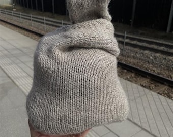 May include: A light gray knitted hat with a hood. The hat is made of a thick, textured yarn and has a folded brim. The hood is attached to the hat and is designed to cover the head and neck. The hat is held in a hand.