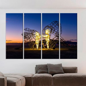 May include: A three-panel canvas print featuring a glowing metal sculpture of two figures standing inside a larger, abstract metal structure. The sculpture is set against a backdrop of a purple and orange sunset sky.