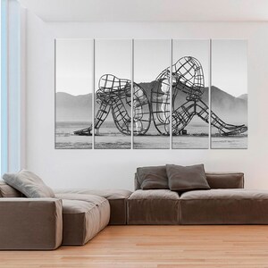 May include: Black and white photograph of a large metal sculpture of two figures intertwined. The sculpture is in a desert landscape.