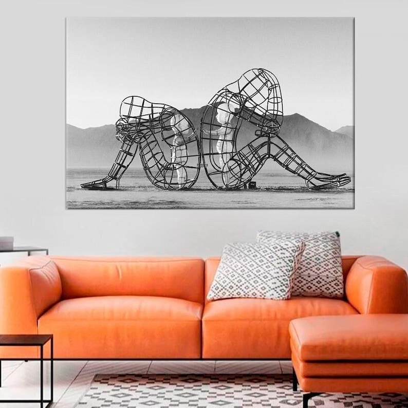 May include: Black and white photograph of a metal sculpture of two figures, one larger than the other, sitting back to back. The figures are made of wire mesh and are posed in a contemplative manner. The sculpture is set against a backdrop of mountains.