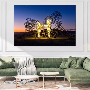 May include: A large metal sculpture of two figures standing in a desert landscape at dusk. The figures are illuminated from within, casting a warm glow against the dark sky.