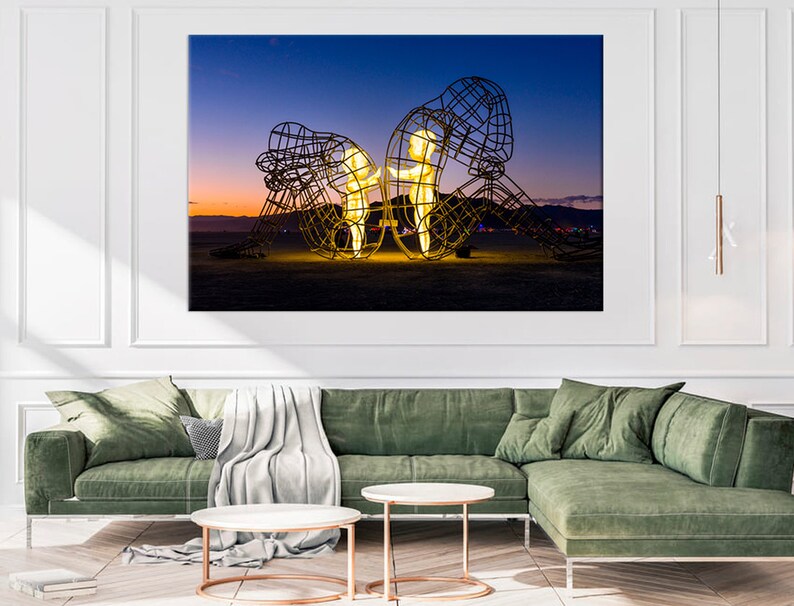 May include: A large metal sculpture of two figures standing in a desert landscape at dusk. The figures are illuminated from within, casting a warm glow against the dark sky.
