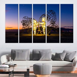 May include: A multi-panel canvas print featuring a large metal sculpture of two figures standing inside a cage-like structure. The sculpture is illuminated from within, casting a warm glow against the backdrop of a desert landscape at sunset.