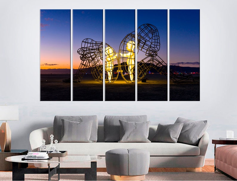 May include: A multi-panel canvas print featuring a large metal sculpture of two figures standing inside a cage-like structure. The sculpture is illuminated from within, casting a warm glow against the backdrop of a desert landscape at sunset.