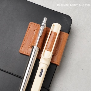 Puede incluir: Un portalápices de cuero color canela sujeto a un cuaderno negro, con dos bucles que sostienen un bolígrafo de metal plateado y uno color crema. El texto Dual Size: 12 mm & 14 mm indica que el soporte admite bolígrafos de estos tamaños.