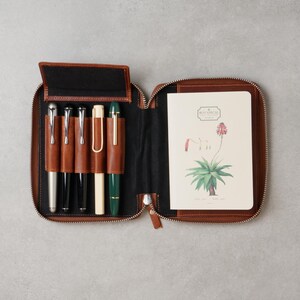 May include: A brown leather zippered case containing five pens in individual slots and a notebook with a botanical illustration of a plant and the text "BOTANICAL" on the cover. The interior of the case is lined with black fabric.