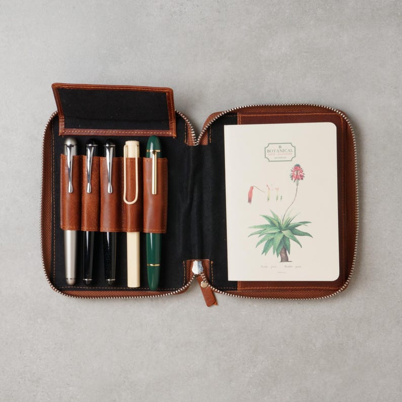May include: A brown leather zippered case containing five pens in individual slots and a notebook with a botanical illustration of a plant and the text "BOTANICAL" on the cover. The interior of the case is lined with black fabric.