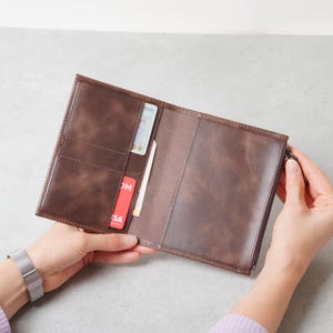 May include: A brown leather passport holder with a zippered compartment on the right side. The interior features multiple vertical card slots on the left side, currently holding a red card and a white identification card.