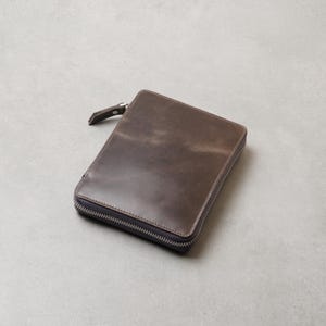 May include: A rectangular dark brown leather case with a smooth, matte finish and a silver-toned metal zipper closure that extends along three sides. The case features a matching leather zipper pull tab attached to the metal slider mechanism.