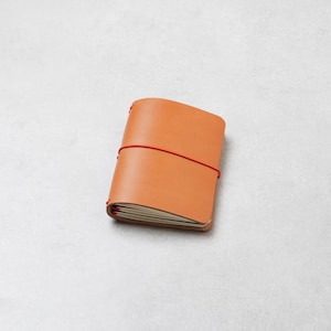 May include: A tan leather notebook cover featuring a smooth, matte finish and rounded corners. The cover is secured by a vibrant red elastic band that wraps horizontally around the centre, holding the internal white paper pages in place.