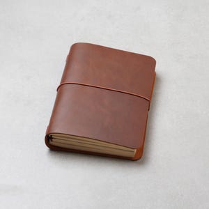Könnte beinhalten: Ein braunes Notizbuch mit einem glatten, einfarbigen Einband, das durch ein dünnes, passendes Gummiband gesichert ist, welches horizontal um die Mitte verläuft. Das Notizbuch enthält mehrere Seiten cremefarbenes Papier.