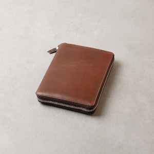 Könnte beinhalten: Ein rechteckiges Etui aus braunem Leder mit glatter Oberfläche. Das Etui verfügt über einen silberfarbenen Metallreißverschluss, der an drei Seiten entlangläuft und an einer kleinen Lederlasche am Reißverschlussschieber oben endet.