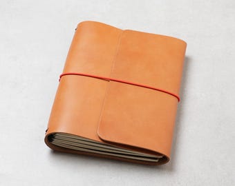 May include: A tan leather notebook cover featuring a smooth, matte finish and a wrap-around design. The cover is secured by a thin, vibrant red elastic band that stretches horizontally across the centre to keep the pages firmly closed.
