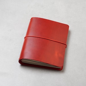 Könnte beinhalten: Ein rotes Notizbuch mit Ledereinband, das mit einem passenden roten Gummiband verschlossen ist. Das rechteckige Journal hat eine glatte, einfarbige Oberfläche und enthält mehrere Seiten weißes Papier am unteren Rand.