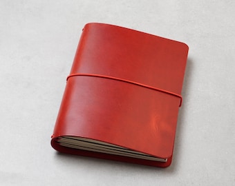 May include: A red leather-covered notebook secured with a matching red elastic band. The rectangular journal features a smooth, solid-coloured exterior and contains multiple pages of white paper visible at the bottom edge of the cover.