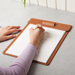 Op de afbeelding: Een bruin lederen klembord met een klep aan de bovenkant bevat een wit plannerblad met gedrukte secties voor datums, prioriteiten en doelen. De plannerpagina heeft een rasterindeling en lijnen voor dagelijkse planning en structuur.