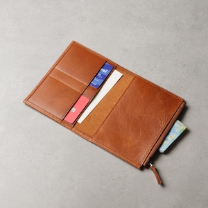 Peut inclure: Portefeuille en cuir marron clair ouvert, avec plusieurs fentes pour cartes, un compartiment zippé et une feuille de papier. Il contient une carte de crédit rouge, une carte bleue avec du texte blanc et de l'argent.