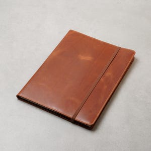 Könnte beinhalten: Ein rechteckiges Notizbuch mit einem glatten, hellbraunen Lederumschlag. Das Notizbuch verfügt über ein passendes schmales Lederband, das diagonal über den vorderen Umschlag verläuft, um die Seiten sicher geschlossen zu halten. Die Kanten sind vernäht.
