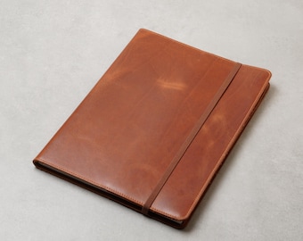 May include: A rectangular notebook with a smooth, tan-colored leather cover. The notebook features a matching thin leather strap that wraps diagonally across the front cover to keep the pages securely closed. The edges are stitched with thread.