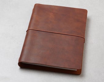 May include: A rectangular notebook with a smooth, medium-brown cover featuring a subtle, textured grain. The notebook is secured by a thin, matching brown elastic band that wraps horizontally around the centre of the front cover.
